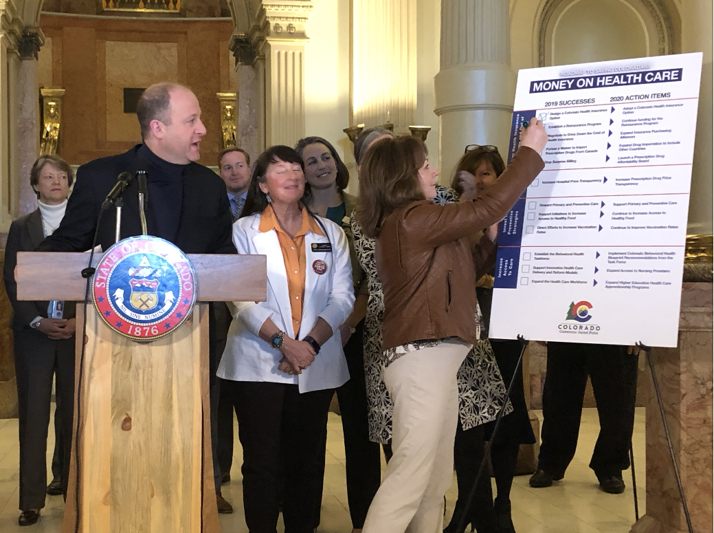 Gov. Jared Polis announces his administration's accomplishments on reducing health care costs, as his lieutenant governor, Dianne Primavera, marks them off during a press conference at the Colorado Capitol on Thursday, Feb. 27, 2020. (Photo by Joey Bunch/Colorado Politics)