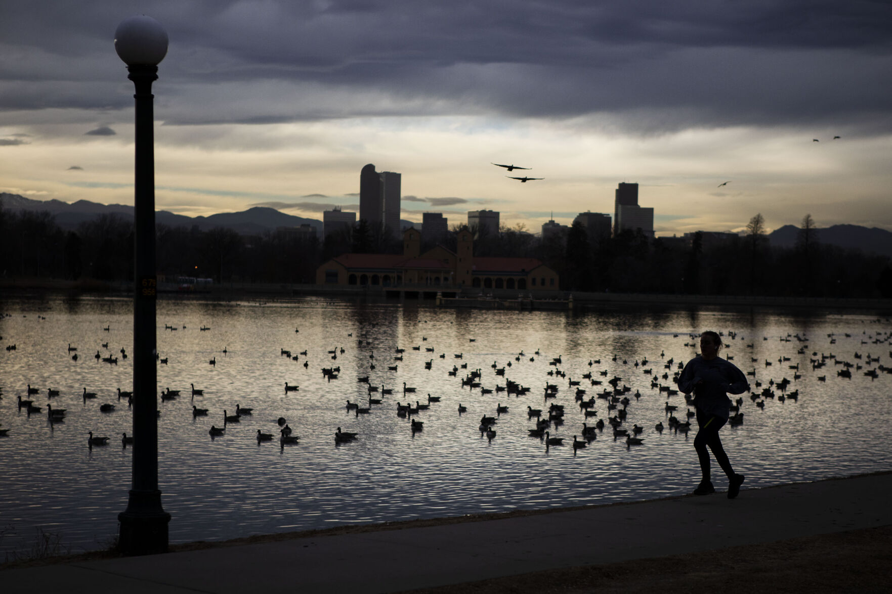Light spills in from behind the Denver skyline as a runner makes their way past Ferril Lake in City Park on Wednesday, Dec. 8, 2021, in Denver, Colo. (Timothy Hurst/The Gazette) (Timothy Hurst)