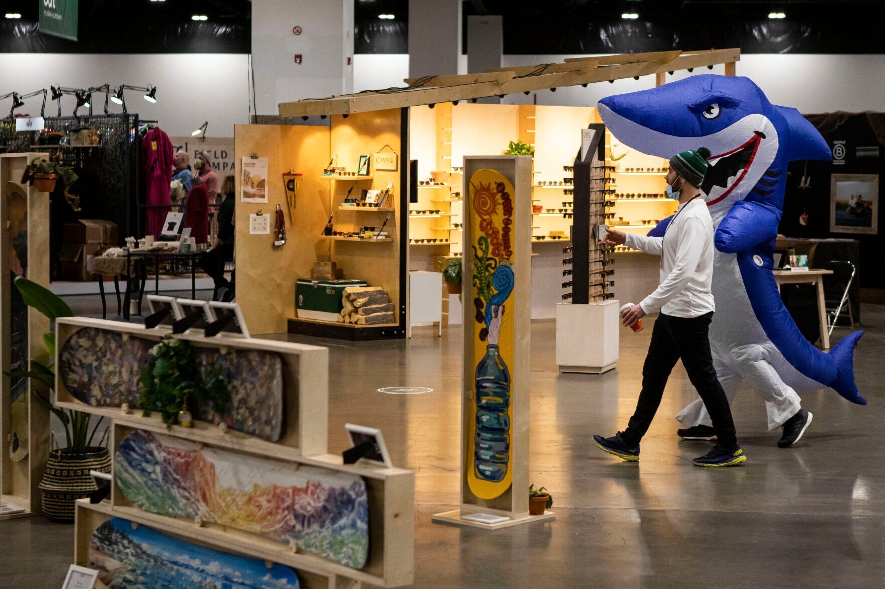 Greg Morrissey, with Mountain Goat Movement, and Tyler Lee with Kavu Inc., dressed in an inflatable shark outfit, walk past a display of snowboards painted by artists during the second day of the Outdoor Retailer Snow Show at the Colorado Convention Center on Thursday, Jan. 27, 2022, in Denver, Colo. (Timothy Hurst/The Gazette) (Timothy Hurst/The Denver Gazette)
