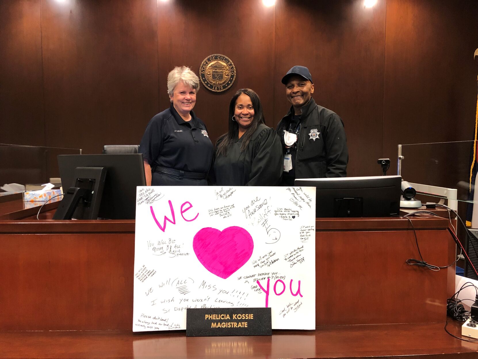 Adams County Magistrate Phelicia Kossie-Tonje's final day in Division R. Bailiffs Jill Colby and Steven Moore are pictured with a sign from deputies and staff at the Adams County Detention Facility. Photo courtesy of Phelicia Kossie-Tonje