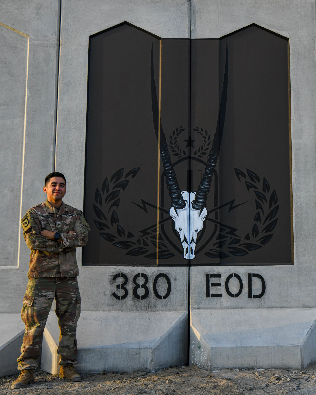 U.S. Army Sgt. Anthony Castillo, a mobility specialist assigned to the 3-157th Field Artillery unit and Colorado National Guard, stands next to a mural he painted for the 380th explosive ordnance disposal flight, September 17, 2022 at Al Dhafra Air Base, United Arab Emirates. The wall features an Arabian Oryx skull (the UAE national animal) combined with the EOD occupational badge – also known as the “Crab.” (U.S. Air Force photo by Tech. Sgt. Jeffrey Grossi) (Tech. Sgt. Jeffrey Grossi)