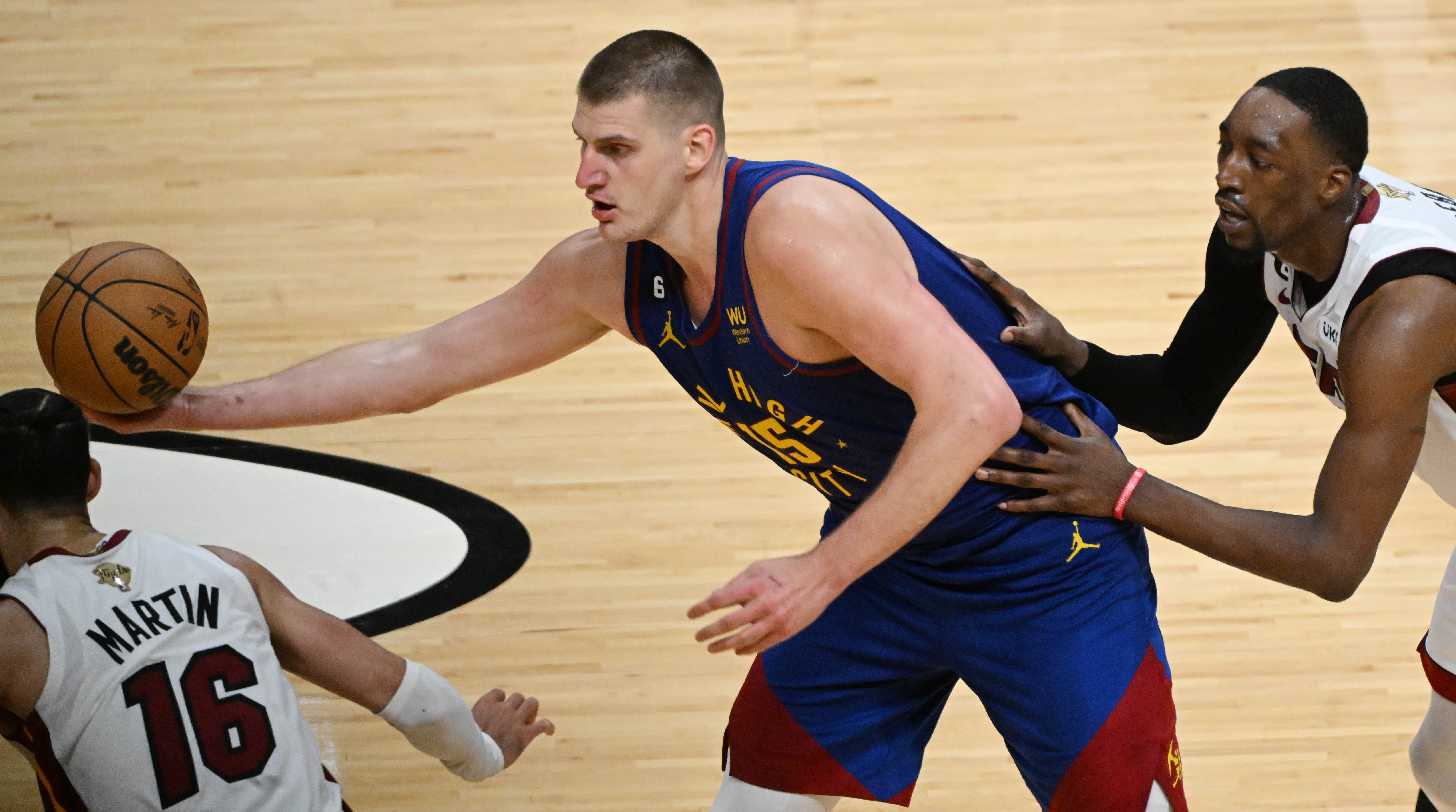 Denver Nuggets center Nikola Jokic (15) hands off the ball while being defended by Miami Heat forward Caleb Martin (16) and Miami Heat center Bam Adebayo (13) during game 3 at Kaseya Center in Miami on Wednesday, June 7, 2023. (Photo by Jerilee Bennett, The Gazette)