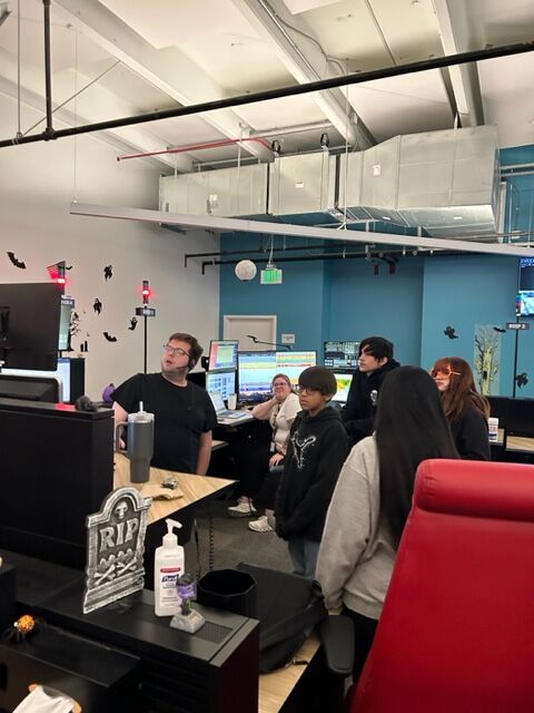 Anthony Lavergne teaches middle schoolers what happens when emergency calls come into a 911 dispatch center. Emergency Communication Technician specialist Kathryn Romero watches from in front of several monitors. (Courtesy: Mike Hess/Denver Police Museum)