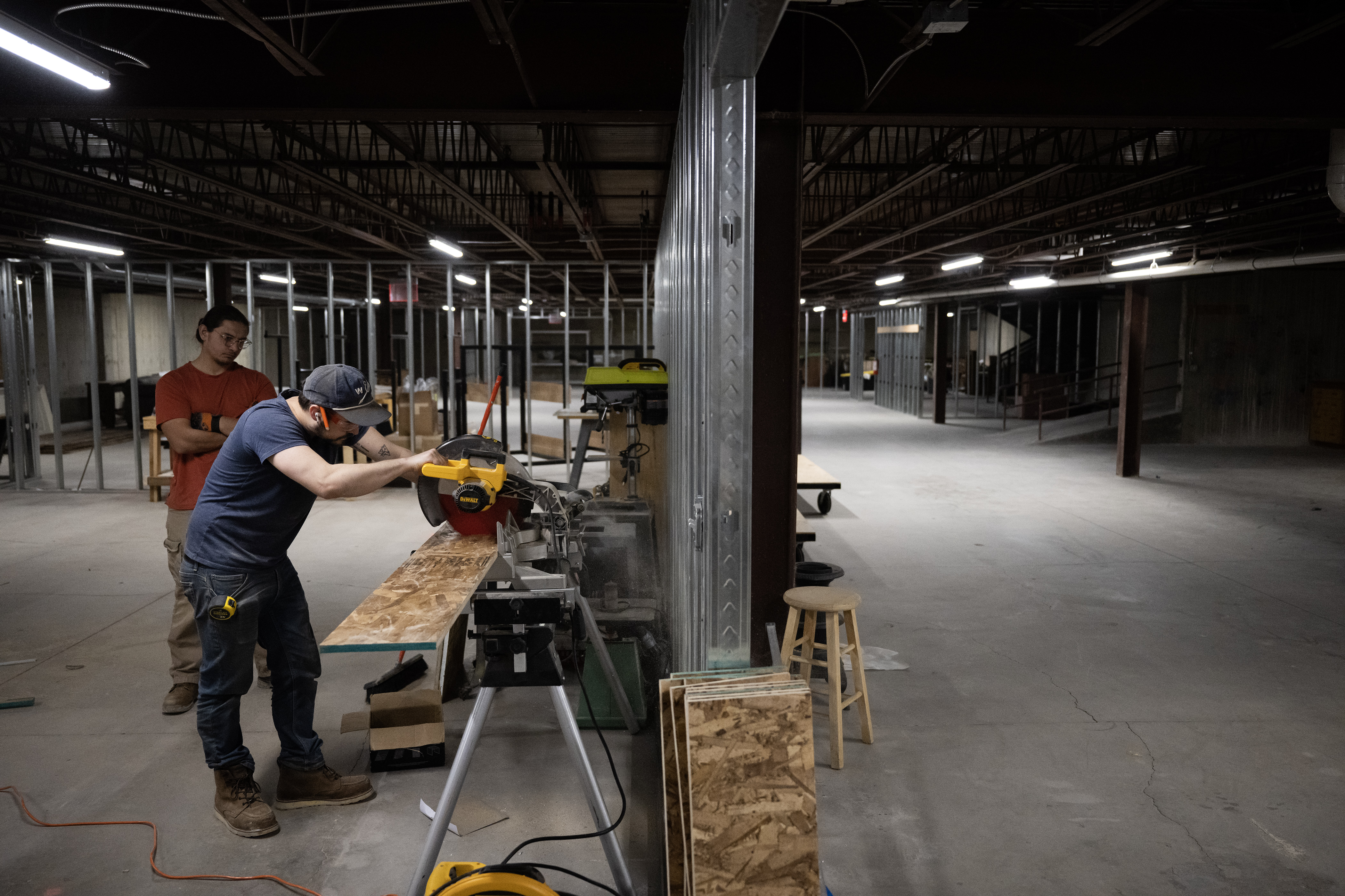 Tanis Gonzales, left, looks on as John Hauser cuts down a length of particleboard in the spacious basement shop for the set of