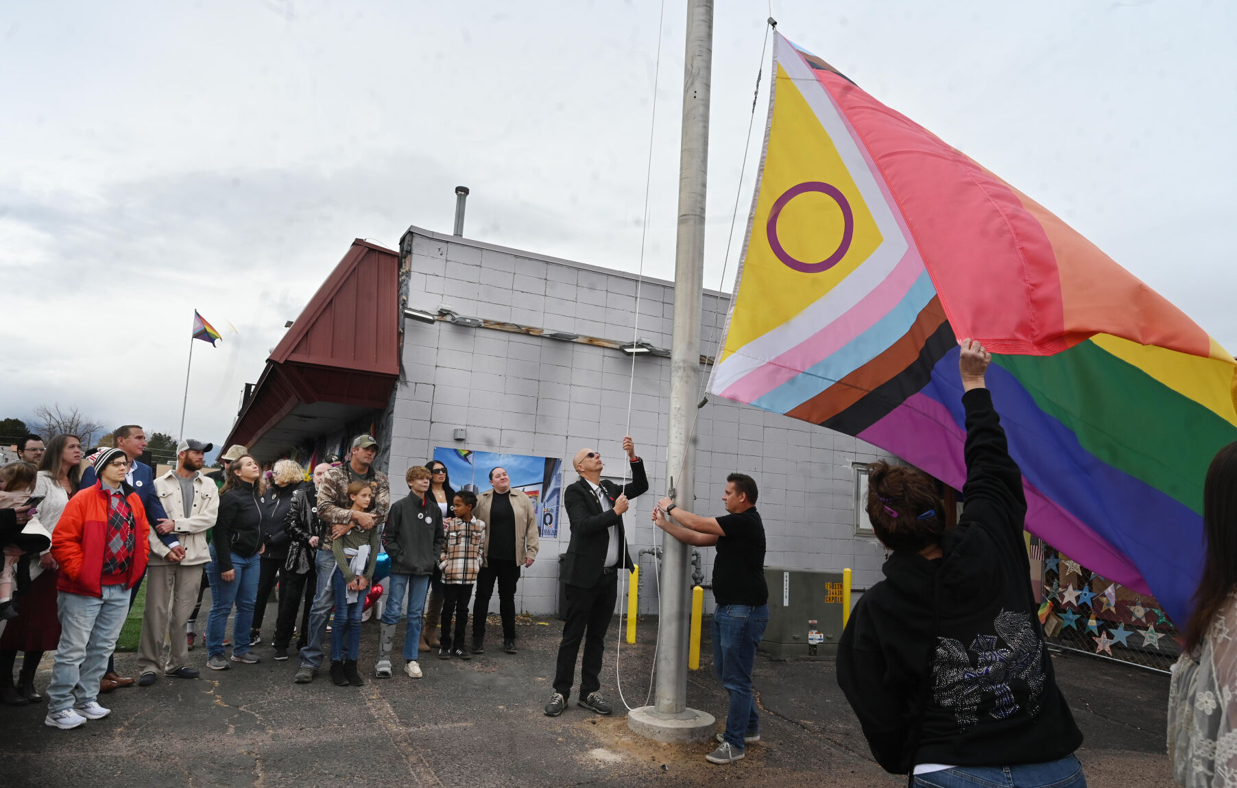Families of victims and survivors of the Club Q shooting gather around a flagpole as a gay pride flag is raised at the site of the shooting. A memorial was held outside of the now closed Club Q, marking the one-year anniversary of the mass shooting on Sunday, Nov. 19, 2023. A year ago, Kelly Loving, Derrick Rump, Ashley Paugh, Raymond Green Vance and Daniel Aston were killed in the nightclub shooting and 17 were injured. (Photo by Jerilee Bennett, The Gazette)