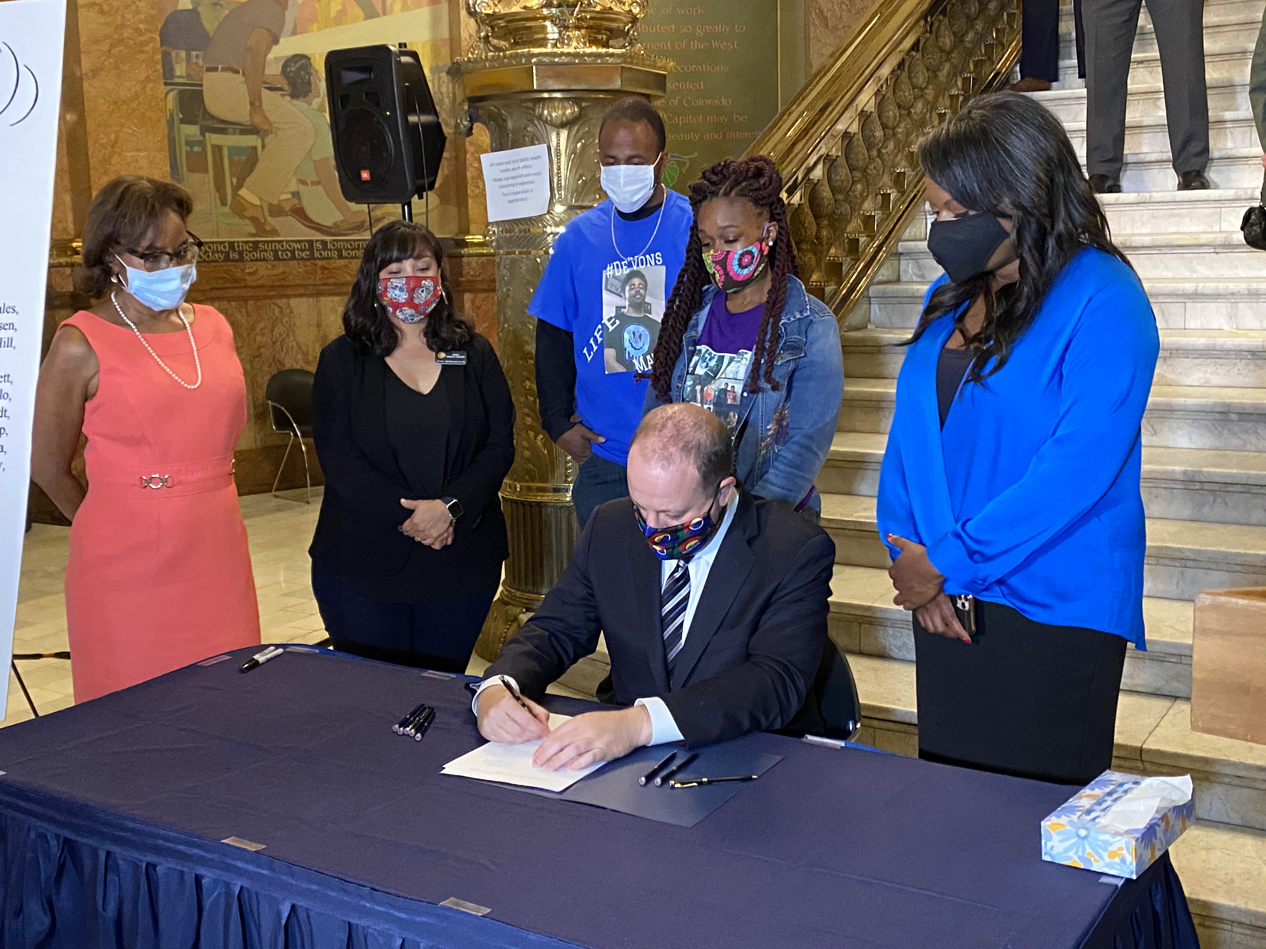 SB217 sponsors (from left) Sen. Rhonda Fields, Rep. Serena Gonzales-Gutierrez and Rep. Leslie Herod (far right) stand with the parents of De'Von Bailey, who was shot in Colorado Springs while he was running away from an officer, while Gov. Jared Polis signs the historic measure. (Carol McKinley, special to Colorado Politics)