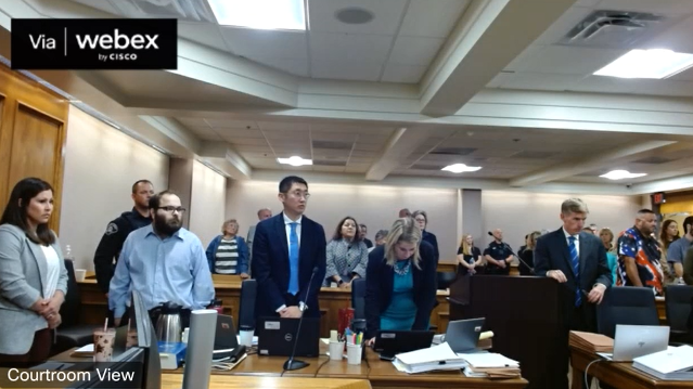 Alleged Boulder mass shooter Ahmad Alissa, standing far left in the blue shirt, in court Monday. Defense attorney Samuel Dunn, of the Colorado Public Defender's Office, is next to him in the blue suit. Boulder District Attorney Michael Dougherty is at the podium. (Screengrab from the 20th Judicial District WebEx feed.)