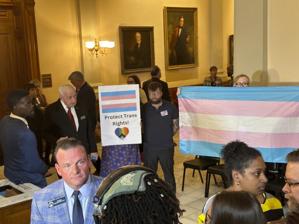 Georgia state Sen. Clint Dixon (R-Buford), left, front, talks to reporters after the passage of a bill that would ban transgender girls from playing high school sports on Tuesday, March, 26, 2024, at the Georgia Capitol in Atlanta. Protesters supporting transgender rights stand behind Dixon (AP Photo/Jeff Amy) (Jeff Amy)