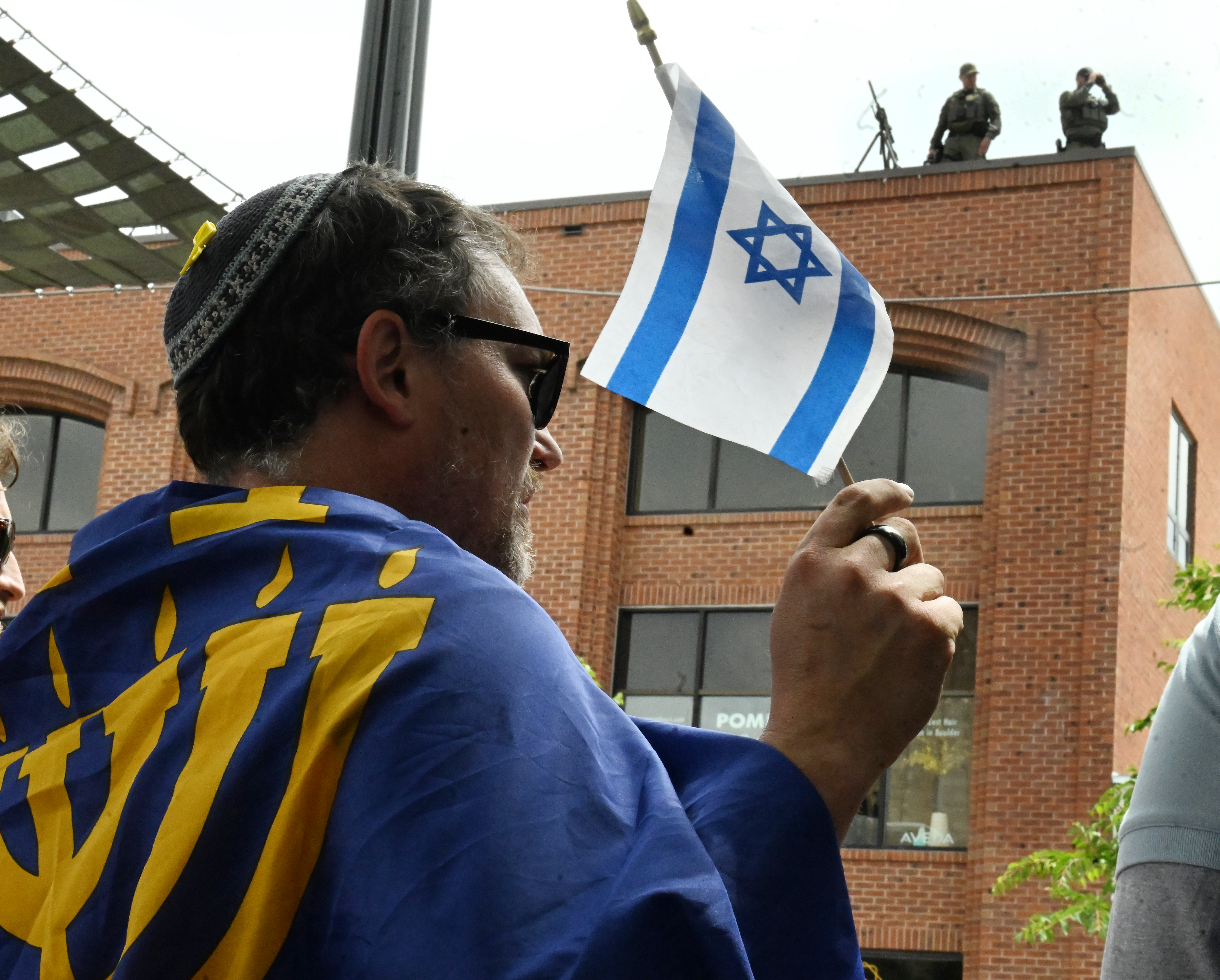 Security with high powered guns are seen on a building top on Pearl Street Mall, as Sam King waves an Israeli flag during a program after the Run For Their Lives walk. They walked in the same location as last week along the Pearl Street Mall on Sunday, June 8, 2025. Last weeks walk ended with a man using makeshift molotov cocktails to set the peaceful protesters on fire. The walk has been held every week in cities around the country to make sure that the hostages in Gaza, that were taken on Oct. 7, 2023, are not forgotten.