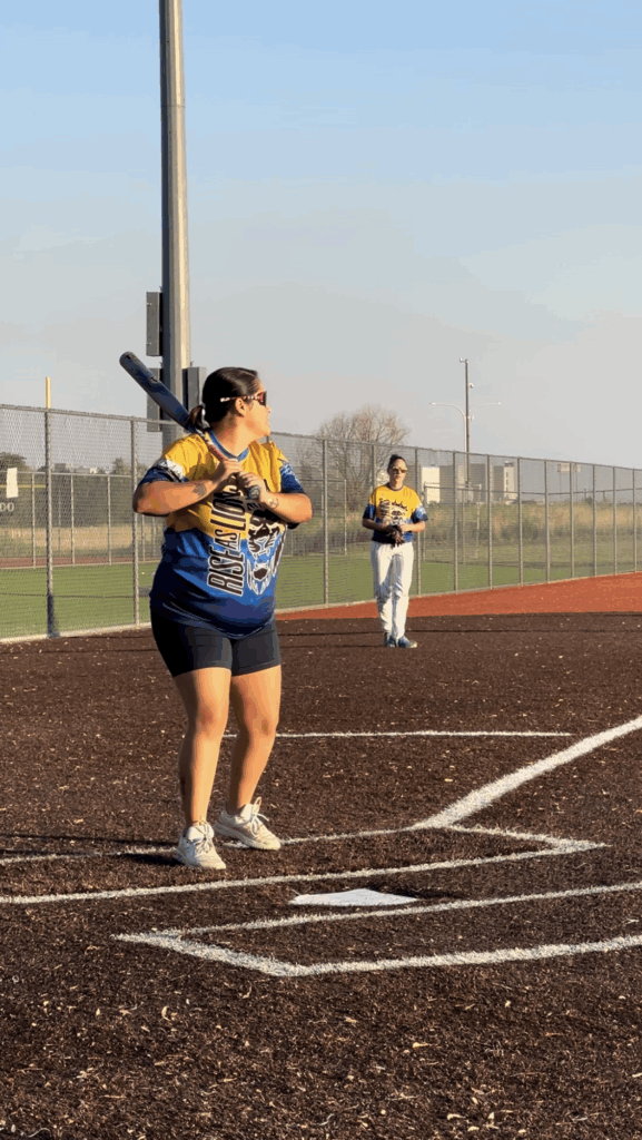 Anna Martinez, outfitted in a yellow and blue Rise as Lions/Recovery Rebels Softball jersey, stands on a baseball field at home plate, readying her bat to swing.