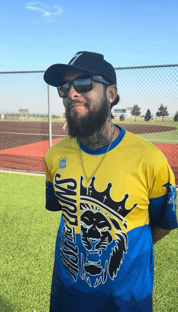 A man in a black baseball cap and black sunglasses stands facing the camera, with his arms folded behind his back. He wears a blue and yellow Rise as Lions/Recovery Rebels Softball jersey.