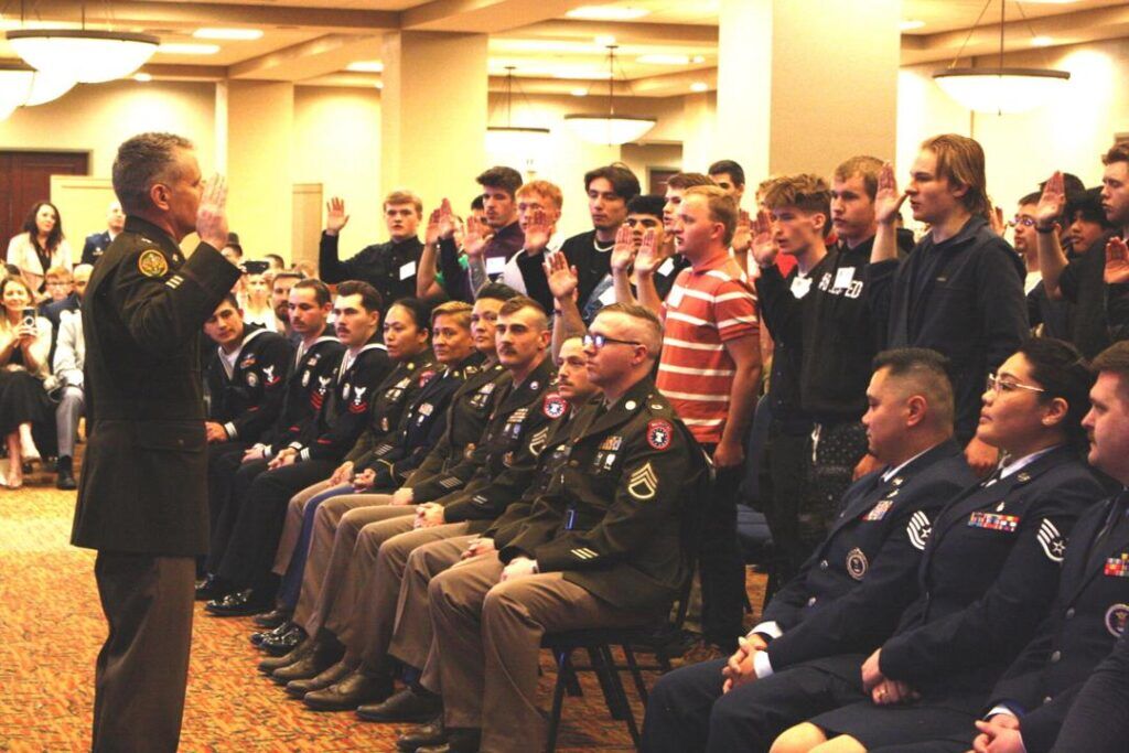 A military officer stands before a room of other officers and high school students.