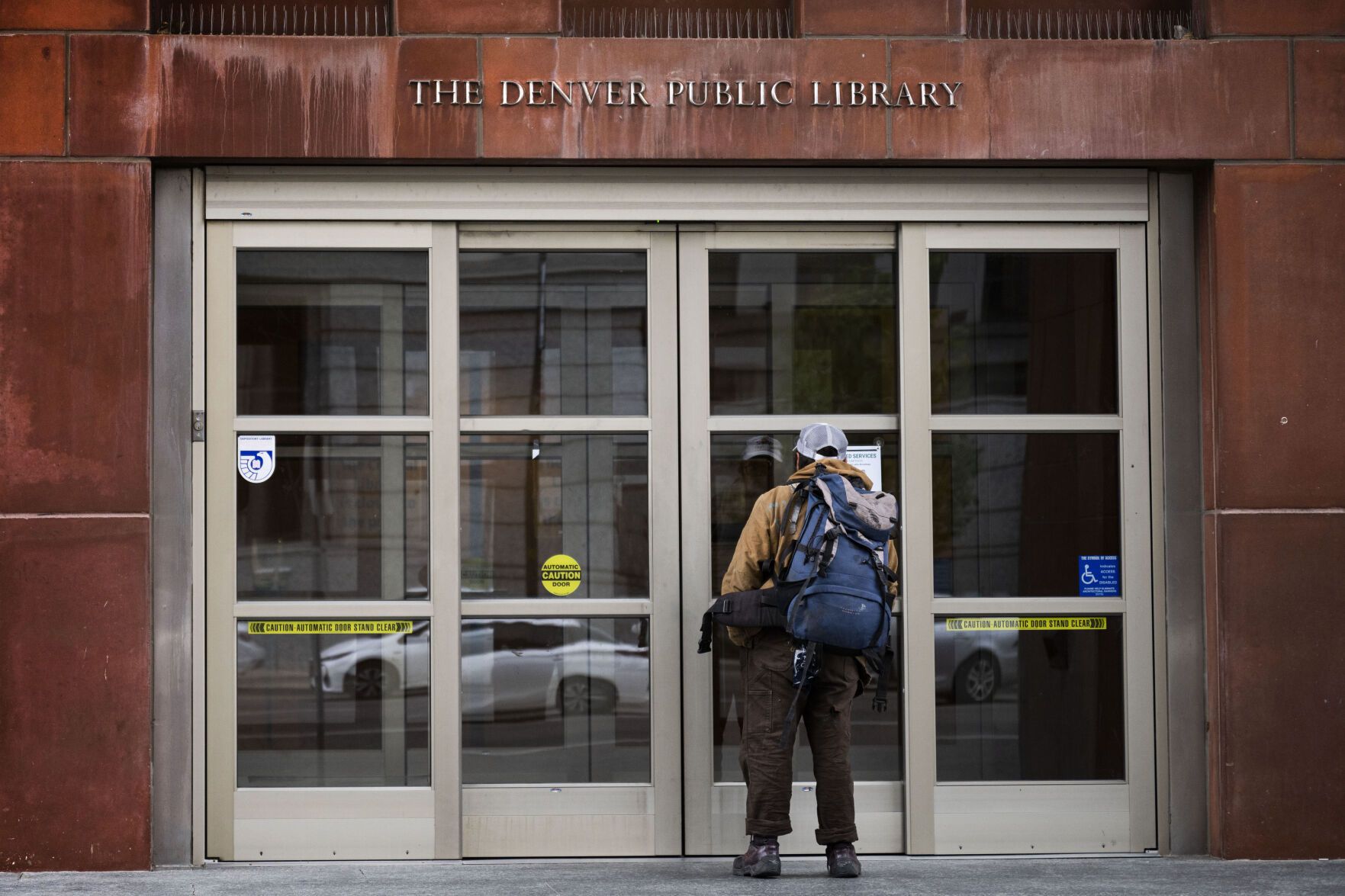 a man with a backpack looks in the front doors of a library building built of red stone