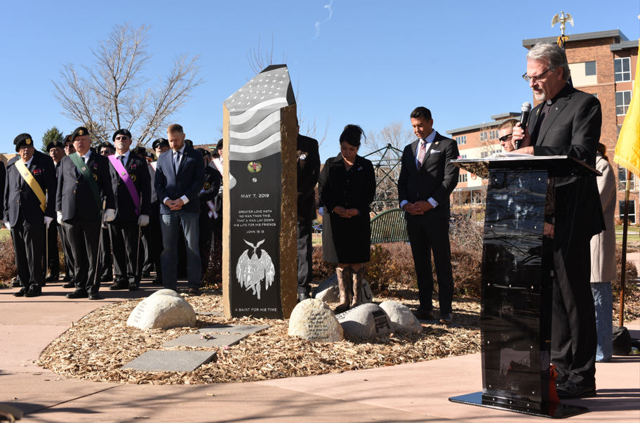 A man stands at a podium in front of a stone memorial with an American flag on top
