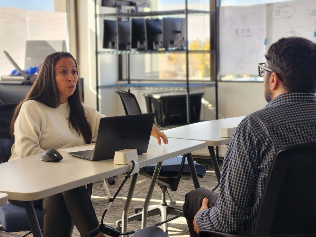 A woman in a white sweater sits behind a test and a laptop and speaks with a dark haired man. 