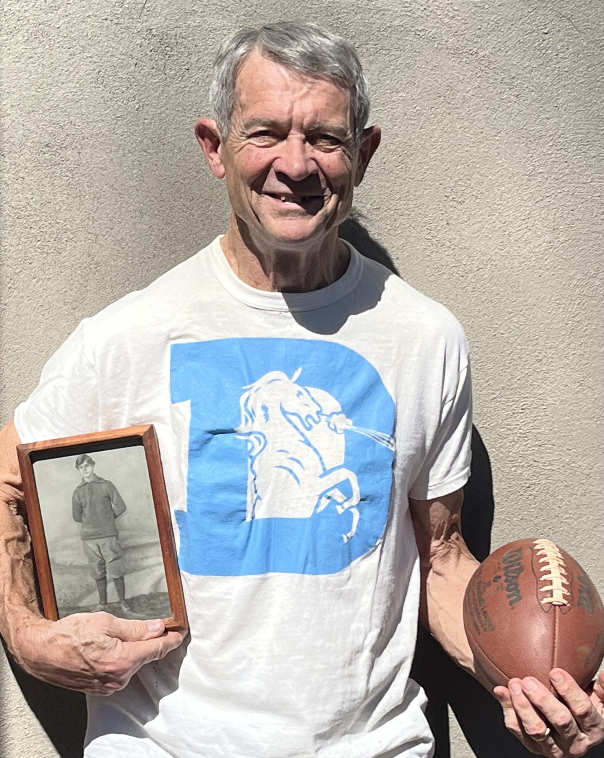 Ken Shelton, a tight end who was a 15th-round pick by the Denver Broncos in 1975 out of Virginia, at his home in Hendersonville, N.C., in March 2025. Shelton elected to soon attend medical school and became a radiologist for 38 years. He is wearing a Broncos T-shirt he received from the team in 1975 while holding a football he got from the team and a photo of his great grandfather N.B. “Bull” Early, who played for Virginia in the 1890s. (Photo courtesy of Ken Shelton) (Photo courtesy of Ken Shelton)