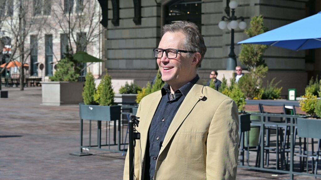 A man in a tan blazer stands at a makeshift podium