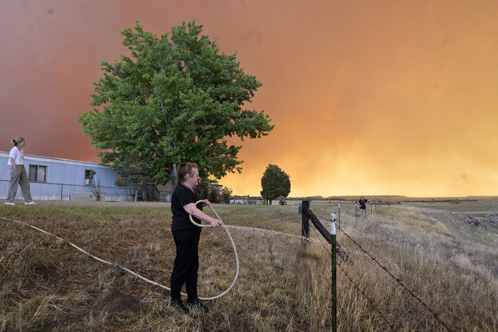 Heather Acavedo uses a garden hose to wet down the grasses around the Mountain Shadows Mobile Estates in hopes of keeping the Aspen Acres fire from reaching the homes in the mobile home park on Monday, June 29, 2026.