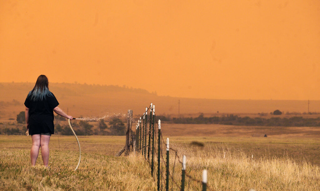 A woman uses a water hose to wet down the grasses around the Mountain Shadows Mobile Estates in hopes of keeping the Aspen Acres fire from reaching the homes in the mobile home park on Monday, June 29, 2026.