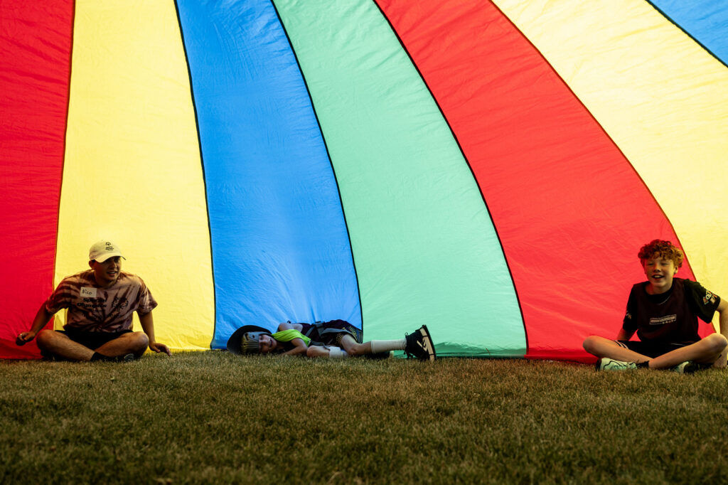 Kids and volunteers sit under a rainbow tarp during a Youth Sports Day clinic for special olympics young athletes on Friday, July 10, 2026. Athletes learned basketball, street hockey, soccer, lacrosse, and dance during the event.