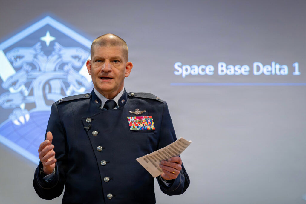 A man in a blue suit speaks in front of a screen that says Space Base Delta 1.